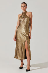 A woman is wearing a gold metallic halter-neck dress that features a keyhole cutout at the neckline and a high slit on the left side, revealing her leg. She is also wearing black ankle-strap high heels.